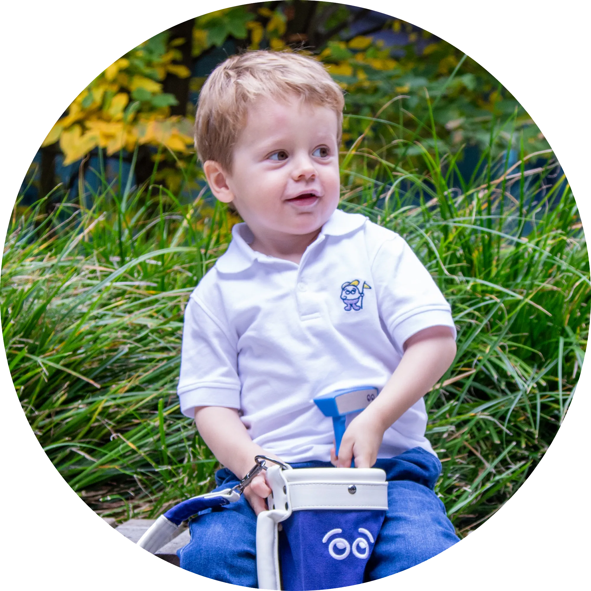 Child in a white polo shirt with a logo, sitting outdoors among greenery