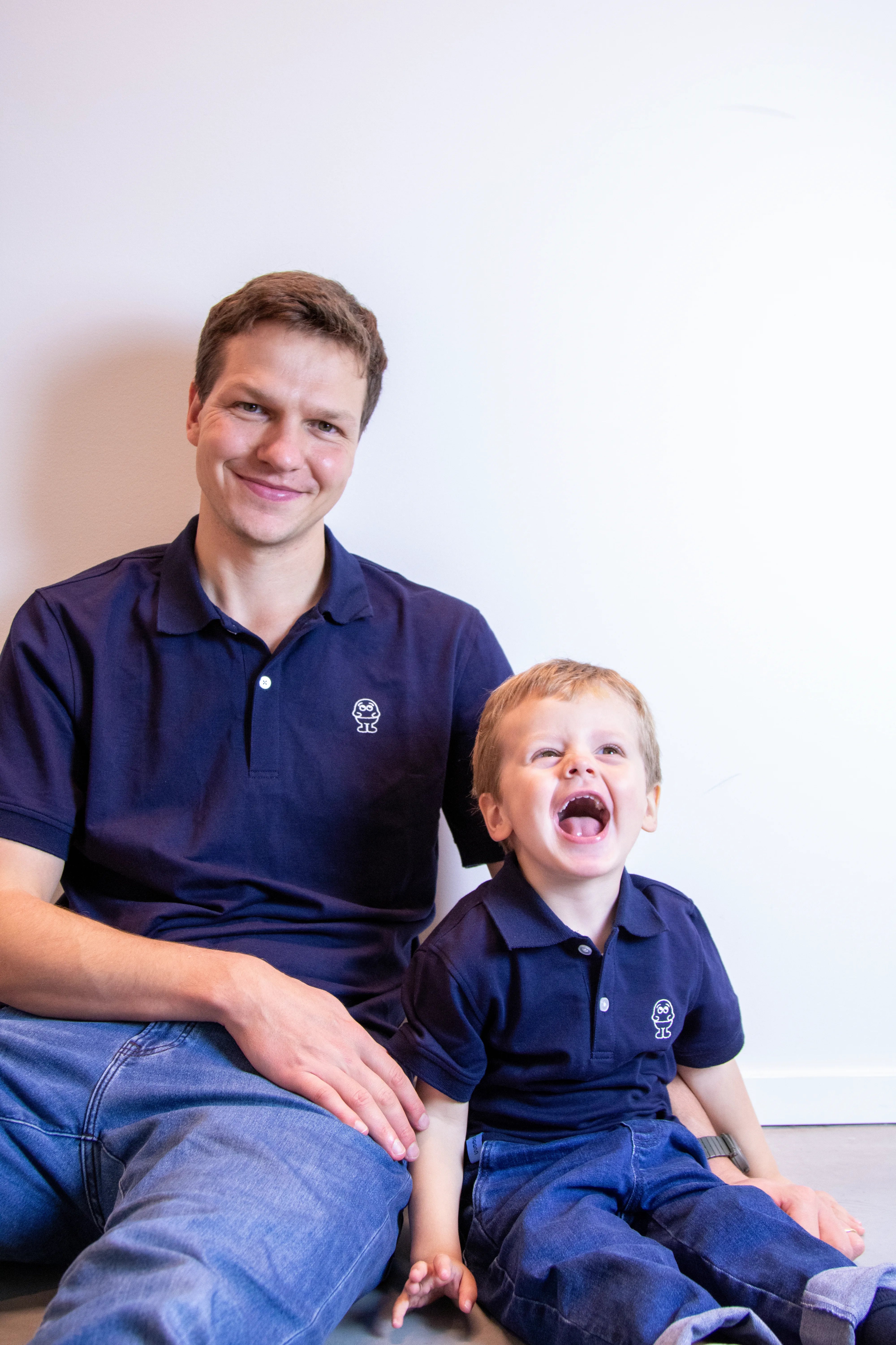 Man and child wearing matching navy blue polo shirts with a logo, sitting against a white wall.