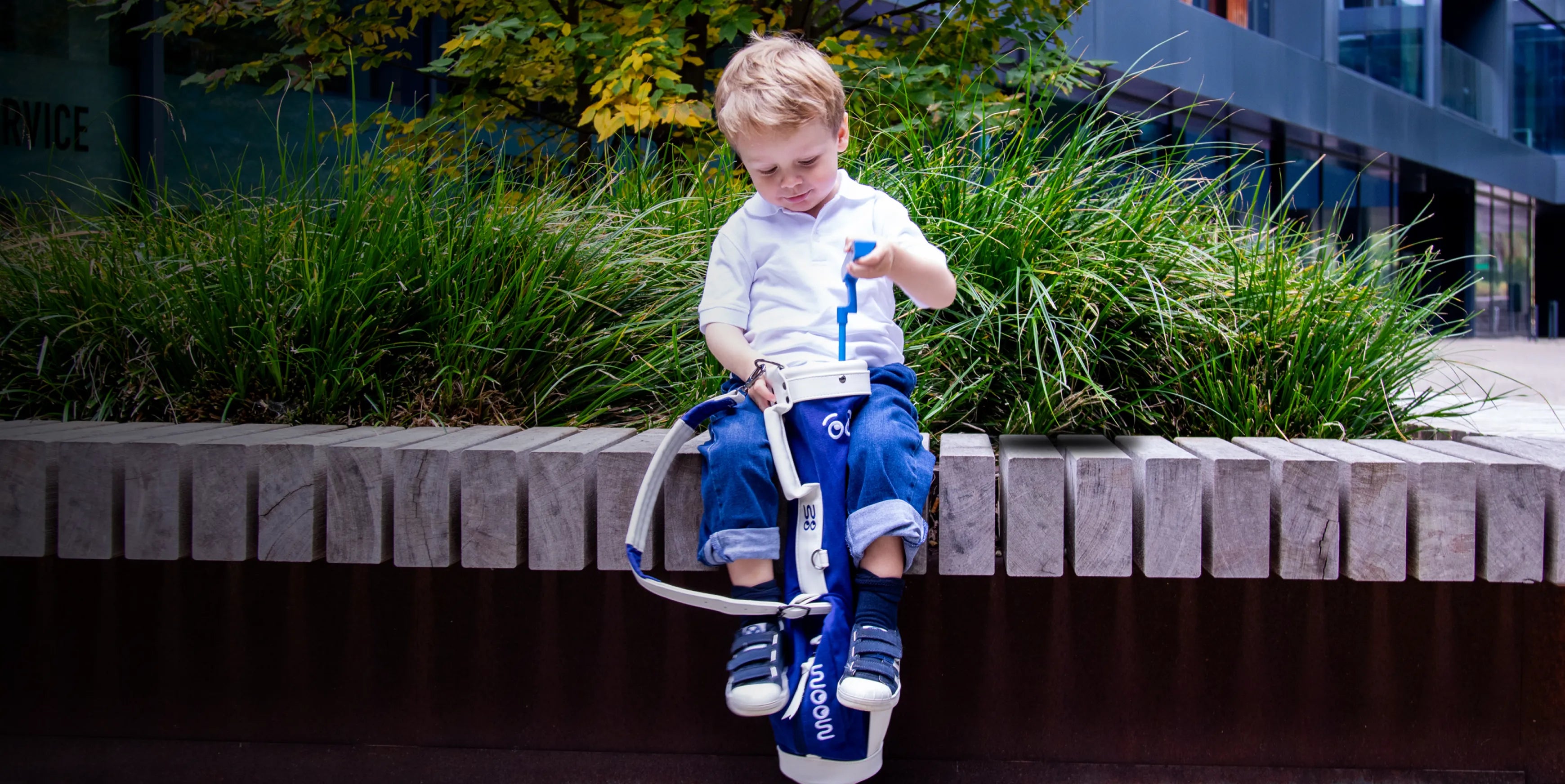 Child sitting on a ledge with a golf bag, surrounded by greenery and a building in the background